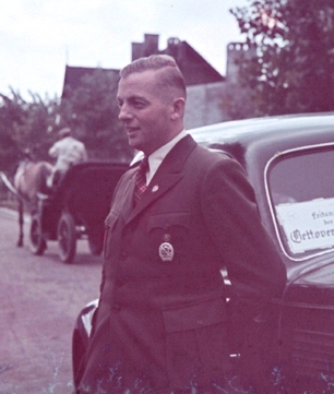 Colored photo of Hans Biebow in his Nazi uniform standing outside of a car with a horse and carriage in the background. 